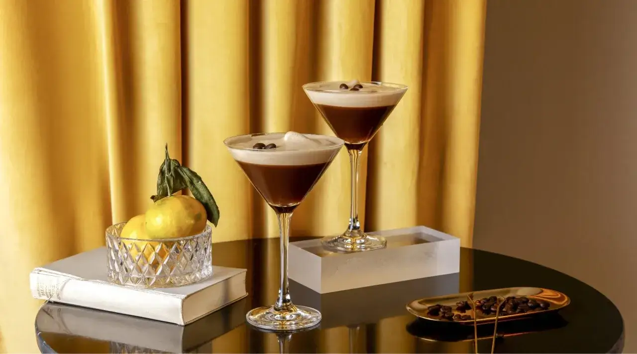 Zwei elegante Gläser mit cremigem Espresso Martini alkoholfrei, garniert mit Kaffeebohnen. Daneben Zitrusfrüchte und Kaffeebohnen auf einem Tablett.