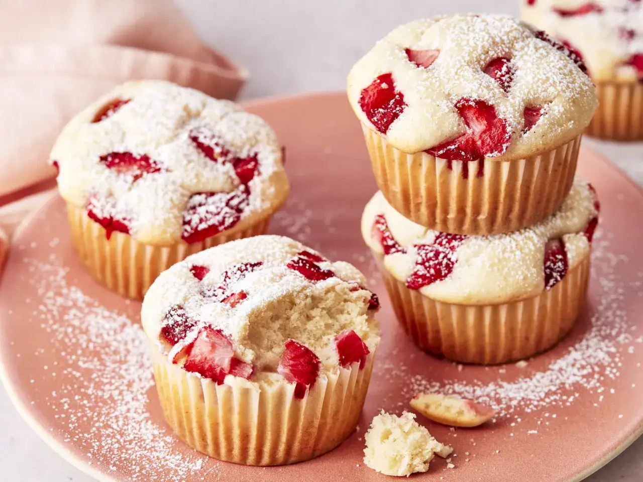 Saftige Erdbeer Muffins, bestäubt mit Puderzucker, liegen auf einem rosafarbenen Teller. Ein Muffin ist angeknabbert.