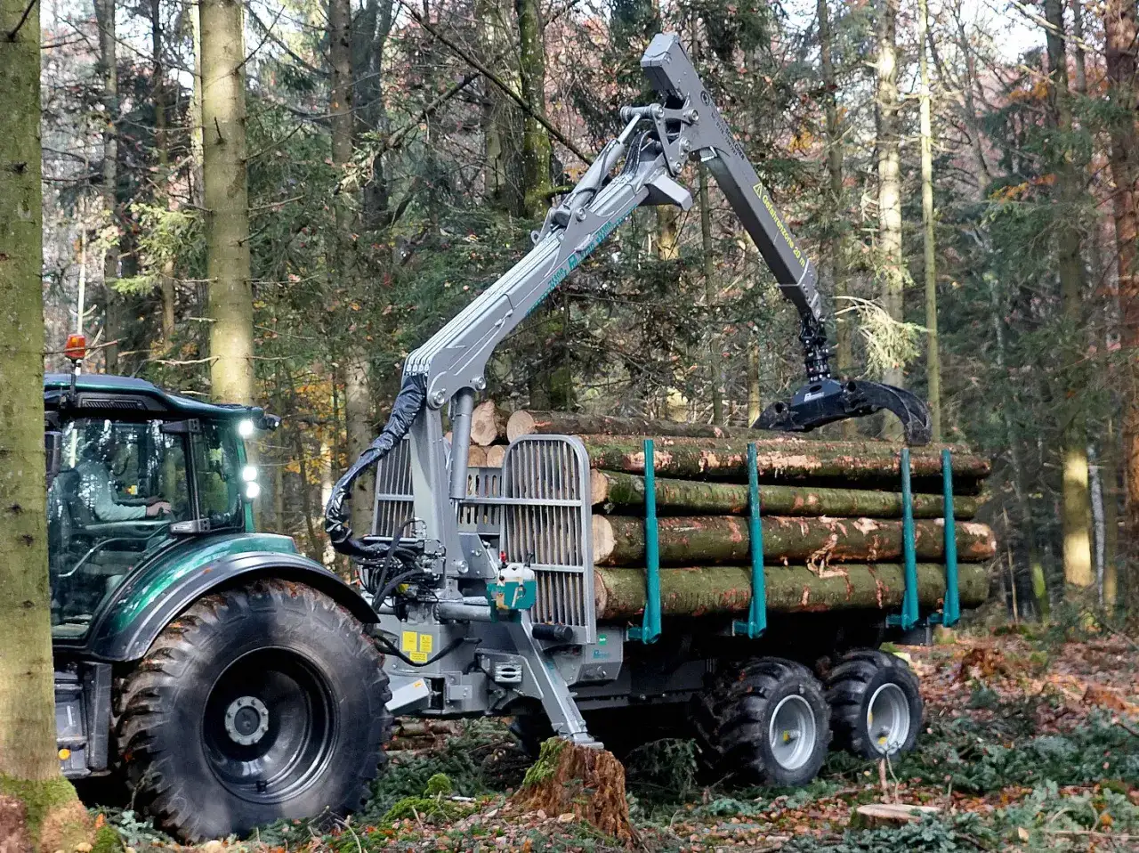 Ein gr&uuml;ner Traktor mit Anh&auml;nger und Ladekran bel&auml;dt Holzst&auml;mme im Wald.