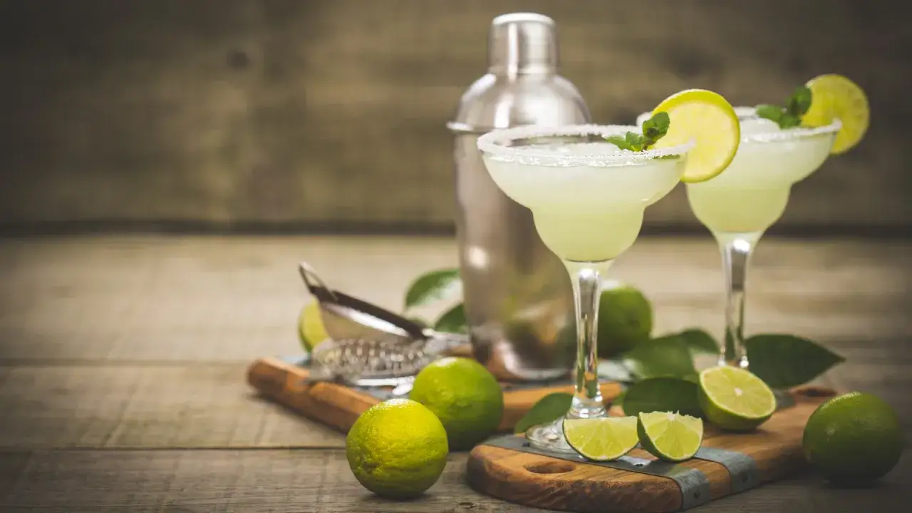 Margarita klasyczna kontra Margarita truskawkowa. Dwa drinki z solą na kieliszkach, limonki i shaker na drewnianym stole.