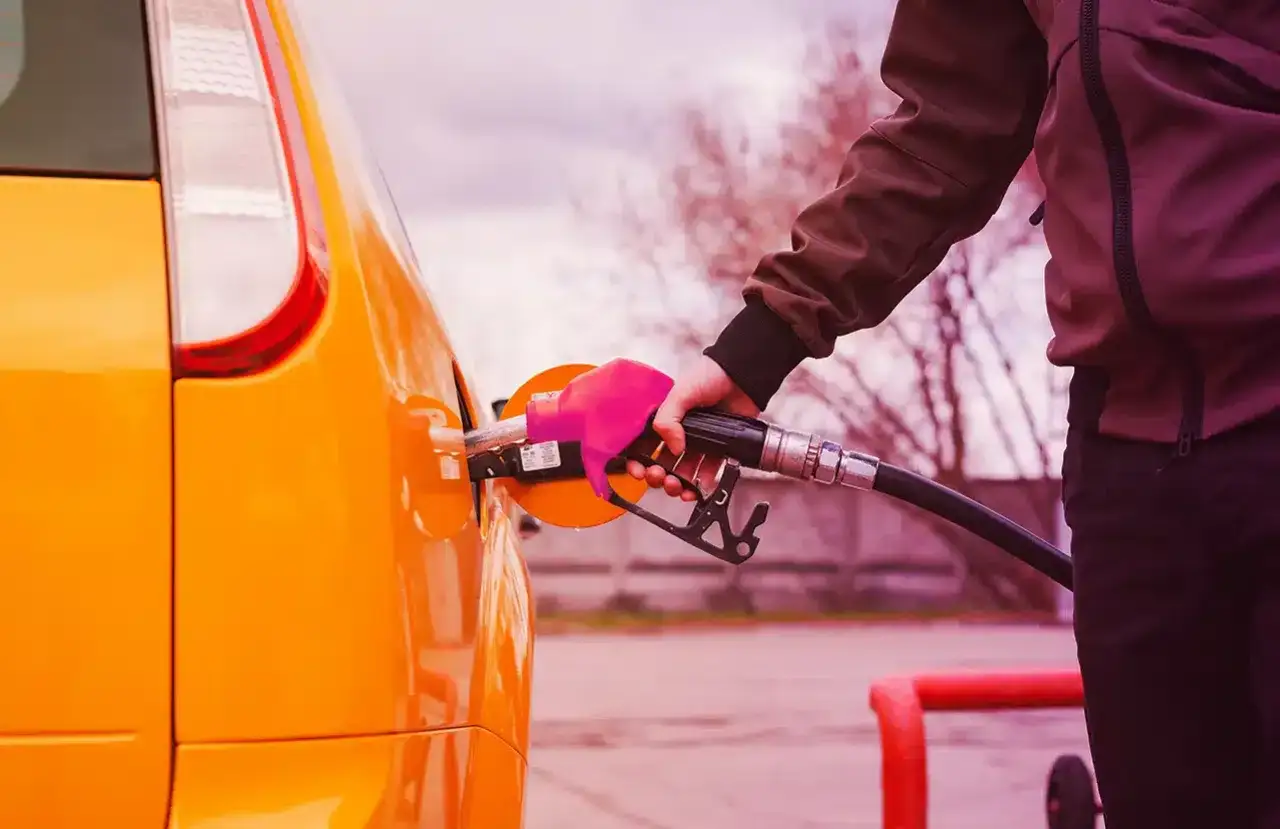 Mano con guanto rosa inserisce la pompa di benzina nel serbatoio di un'auto arancione. Il **consumo auto** è una spesa da considerare.