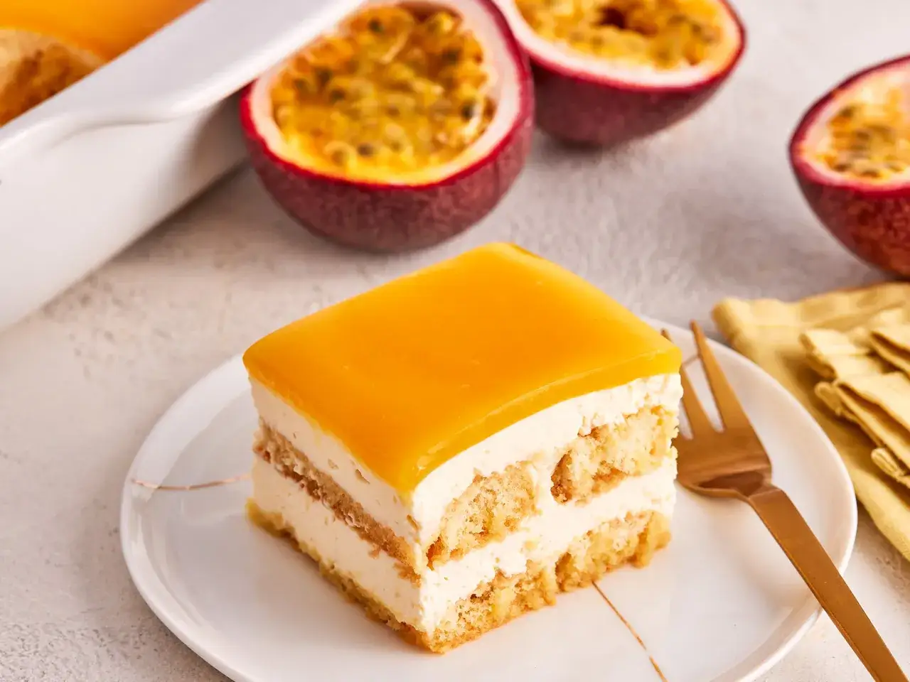 Ein Stück Maracuja Tiramisu mit fruchtiger Geleeschicht und Löffelbiskuits. Daneben liegen aufgeschnittene Passionsfrüchte.