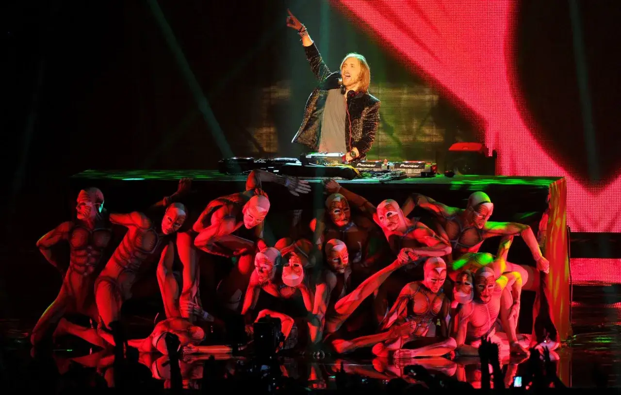 David Guetta na scenie z tancerzami w kostiumach, tworzącymi żywe rzeźby do jego utworów.