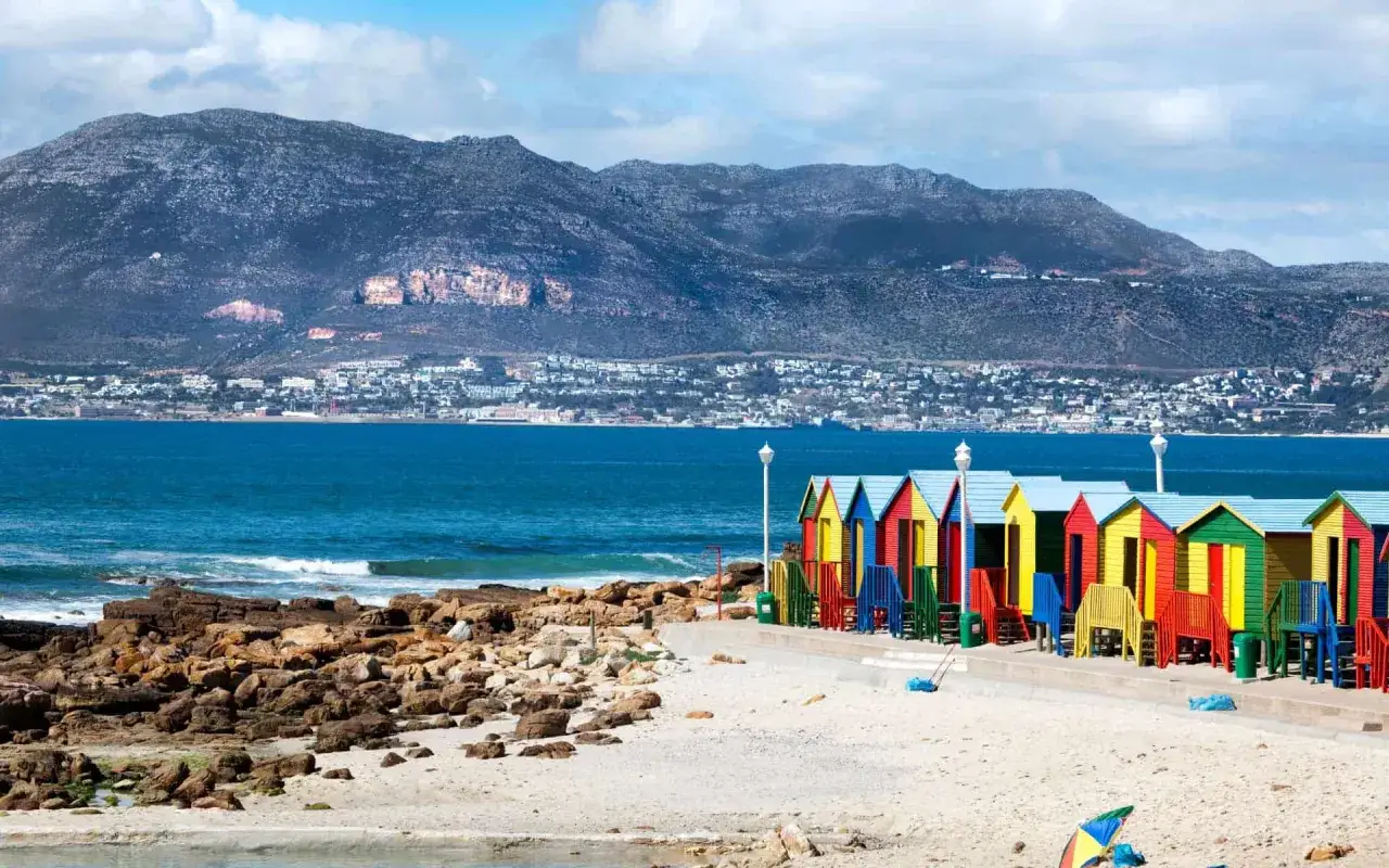Bunte Strandhäuschen am kapstadt strand, mit dem Meer und einer Bergkulisse im Hintergrund.