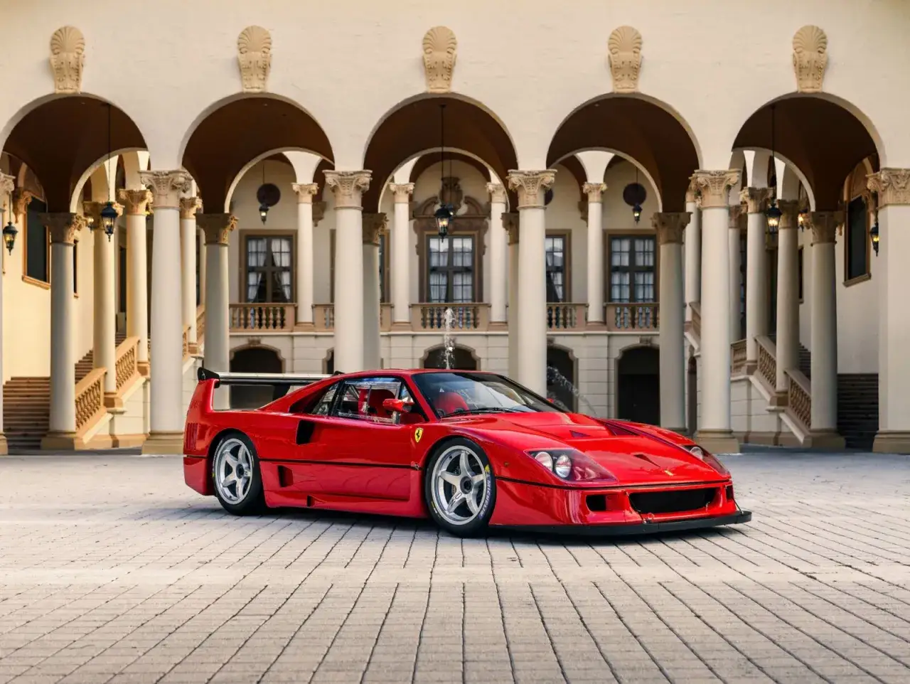 Roter Ferrari F40 LM vor einer prächtigen Villa mit Säulengängen. Ein Traumauto auf gepflastertem Hof.