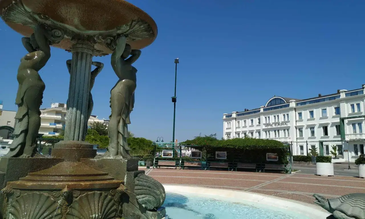 La fontana delle sirene a Cattolica, con le sue figure mitologiche che sorreggono la vasca, si staglia contro il cielo azzurro, con l'Hotel Kursaal sullo sfondo.