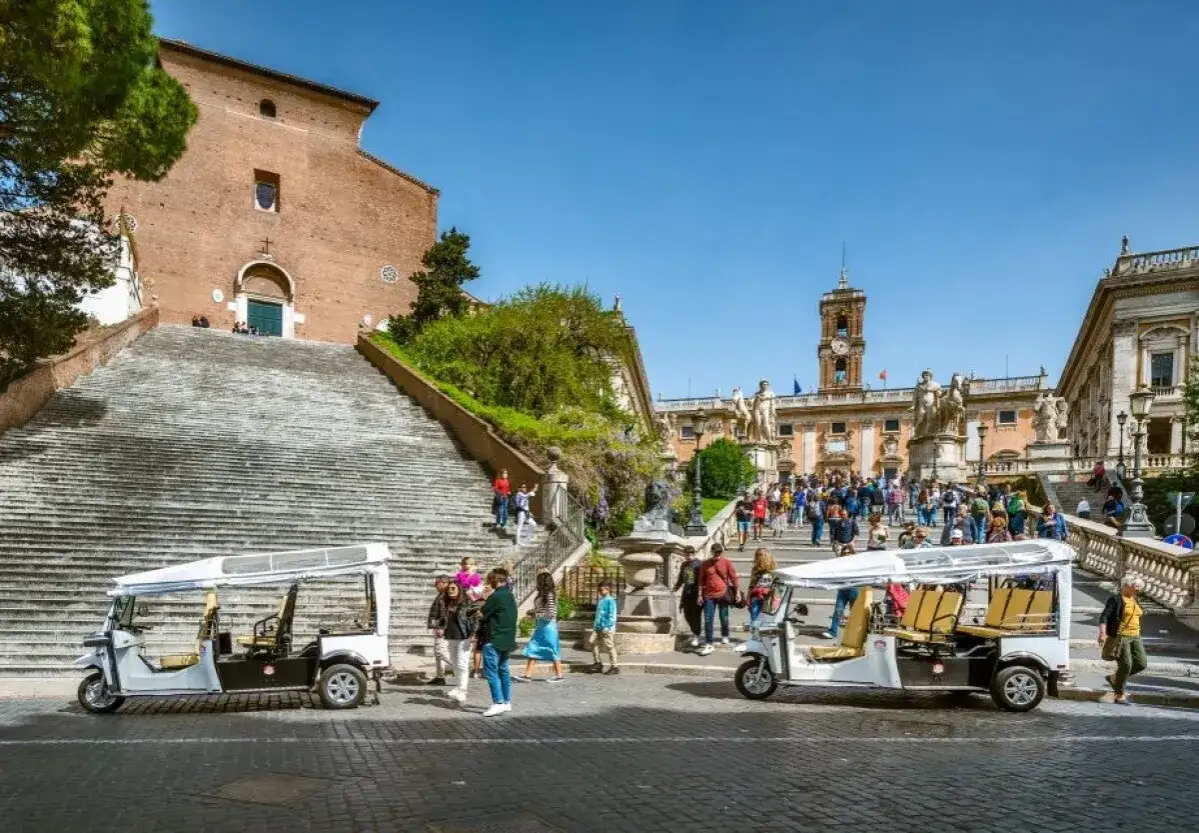 Roma in alta stagione: turisti salgono la scalinata verso il Campidoglio, con golf cart pronti per esplorare la città.