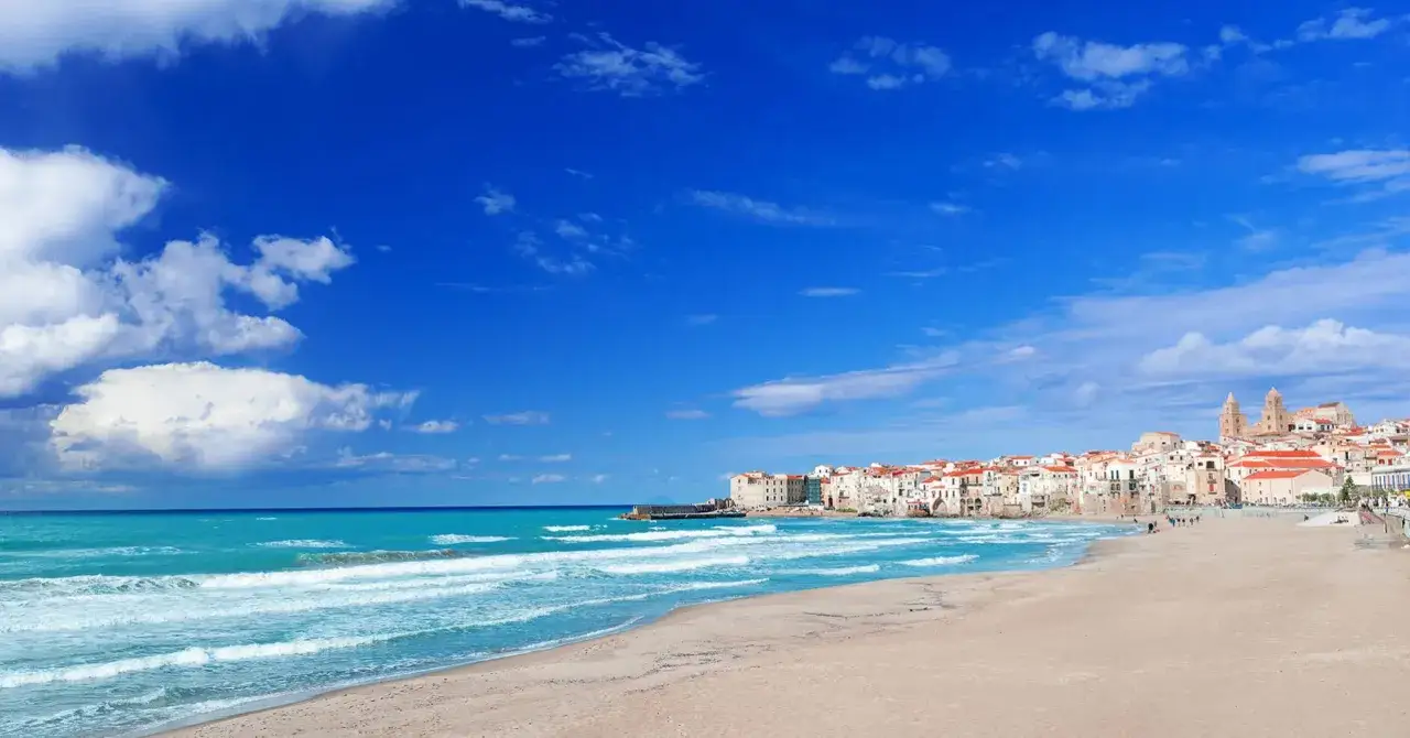Le spiagge di sabbia dorata di Cefalù si estendono sotto un cielo azzurro, con il mare turchese che lambisce la costa.