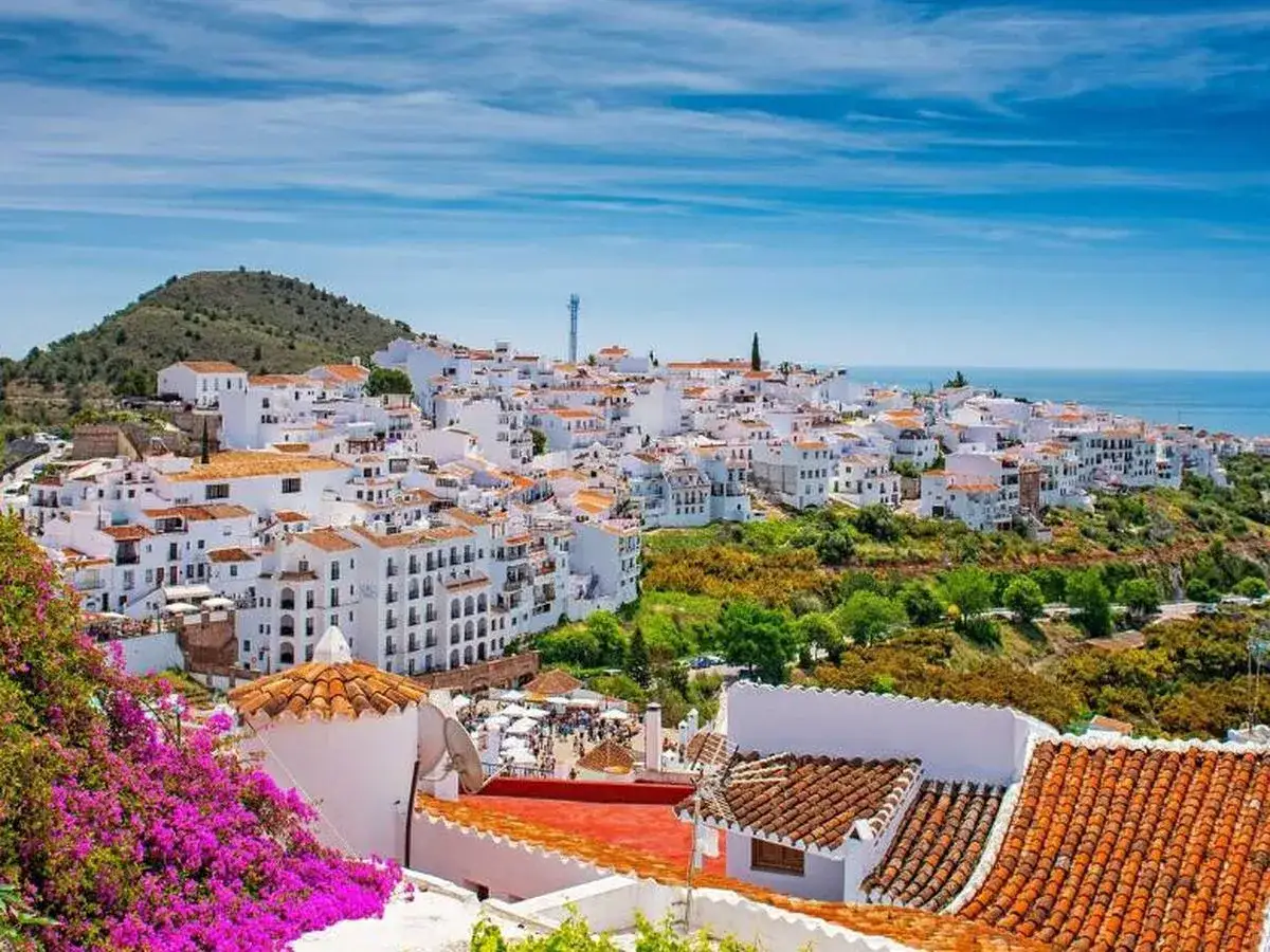 Pueblo blanco en una colina, parte de una ruta andaluza, con el mar al fondo y buganvillas moradas en primer plano.