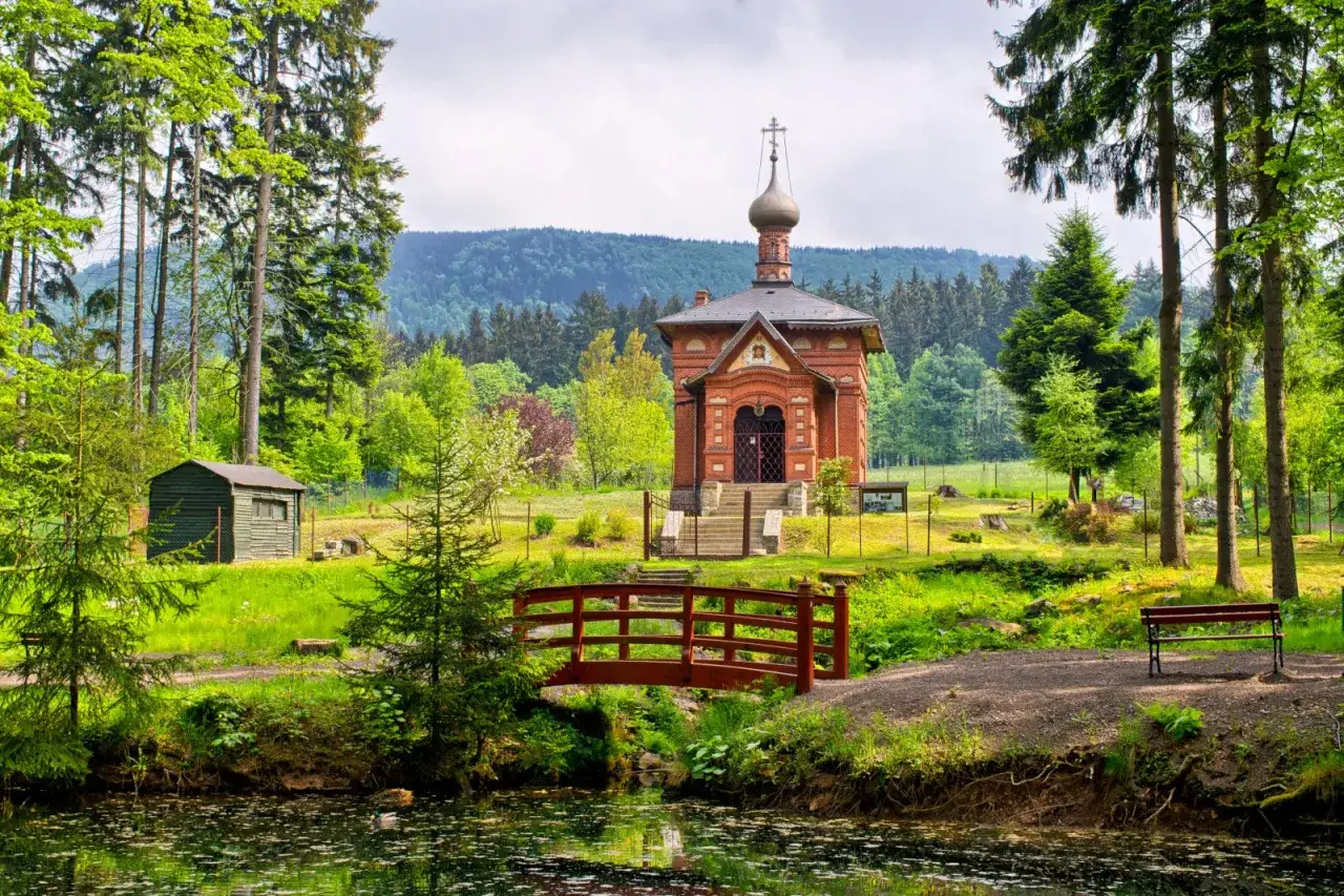 Cerkiewka z czerwoniej cegły z kopułą, otoczona zielenią, z mostkiem i ławką nad wodą. W tle łagodne, zalesione **góry suche**.