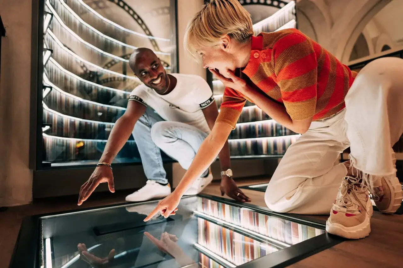Un couple s'&eacute;merveille devant une installation lumineuse dans un mus&eacute;e des paradoxes. Ils pointent du doigt un &eacute;cran interactif au sol, refl&eacute;tant leur joie.
