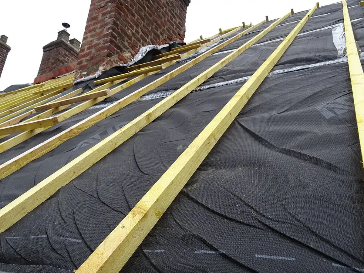 Structure en bois pour une nouvelle ite toiture, avec des chevrons jaunes sur une membrane noire. Des cheminées en briques rouges se dressent au loin.