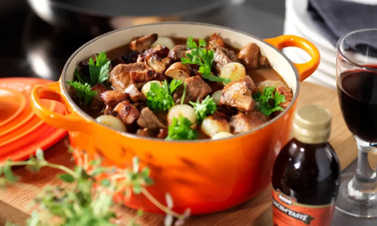 En mustig boeuf bourguignon i orange gryta, med champinjoner, l&ouml;k och persilja. Ett glas r&ouml;dvin bredvid.