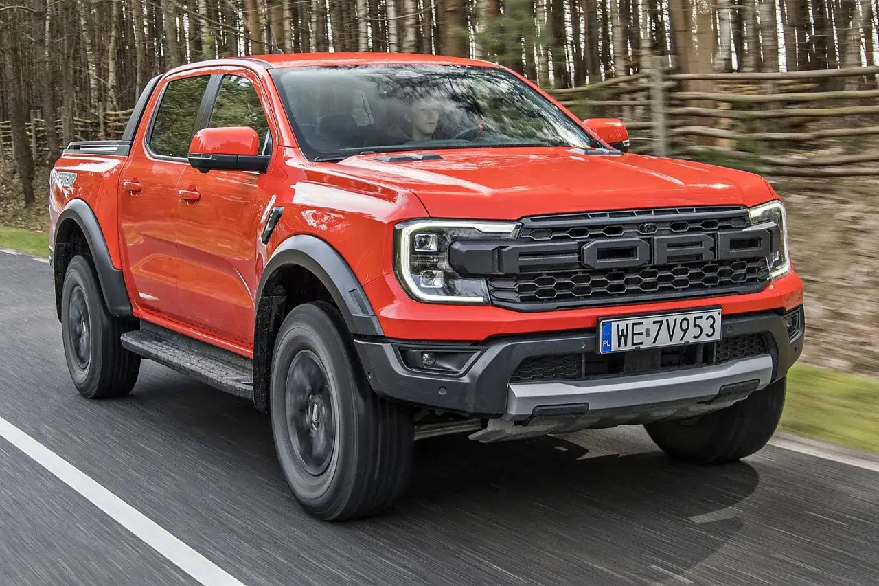 Pomarańczowy Ford Raptor jedzie po drodze w lesie. Sprawdź aktualną ford raptor cena.