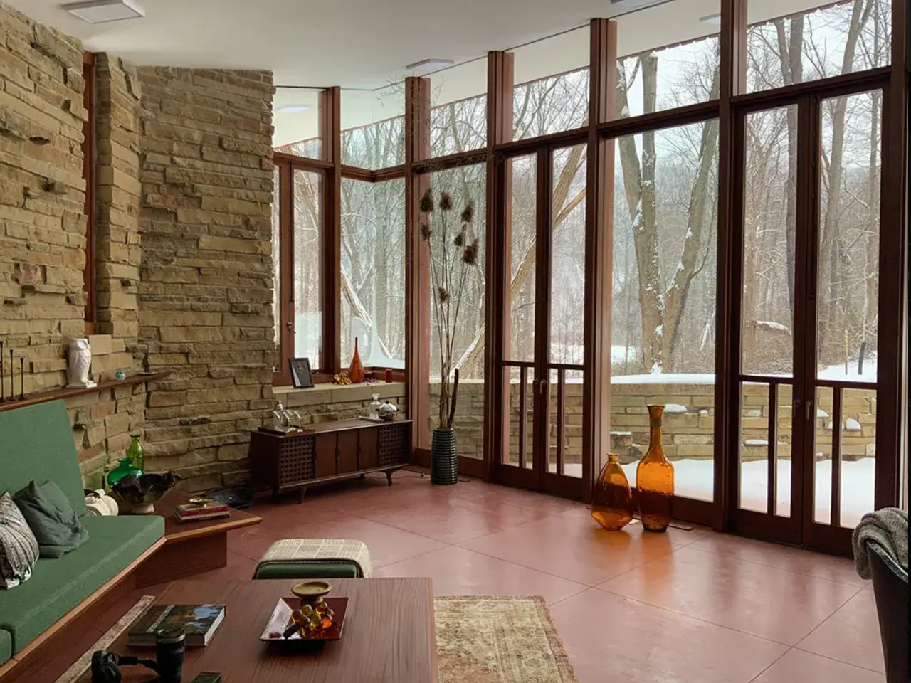Cozy living room in the last Wright house, with stone walls, large windows overlooking a snowy forest, and mid-century furniture.