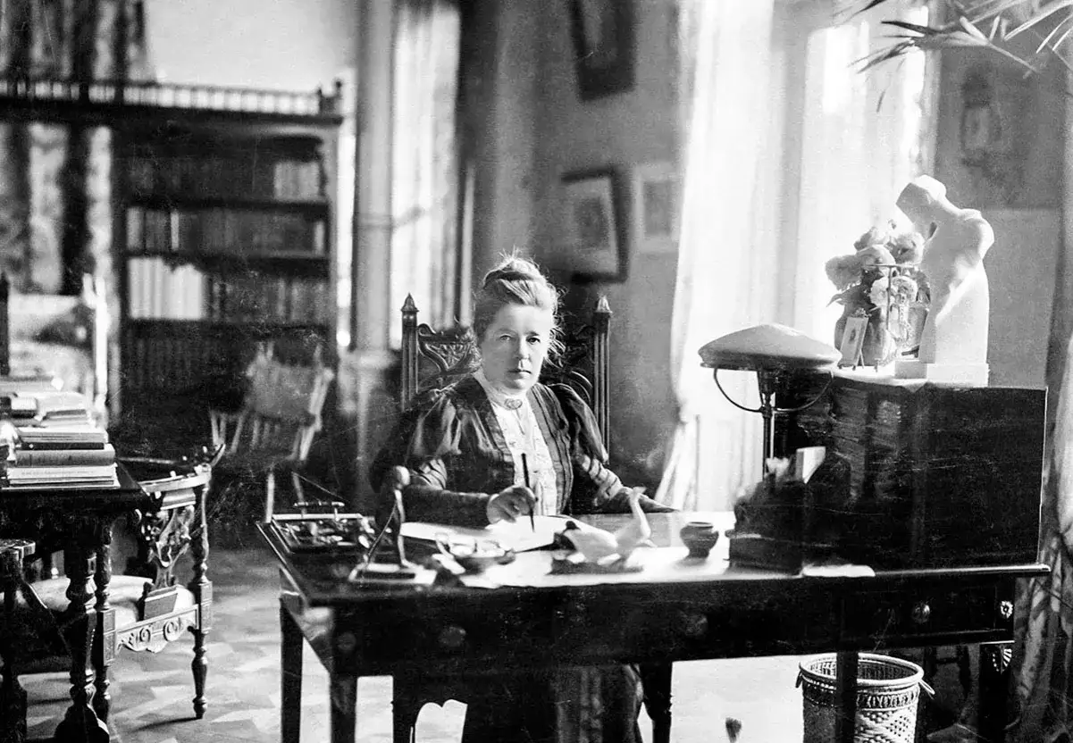 Selma Lagerl&ouml;f sitter vid sitt skrivbord, omgiven av b&ouml;cker och skrivmaterial, redo att skriva sin n&auml;sta ber&auml;ttelse.