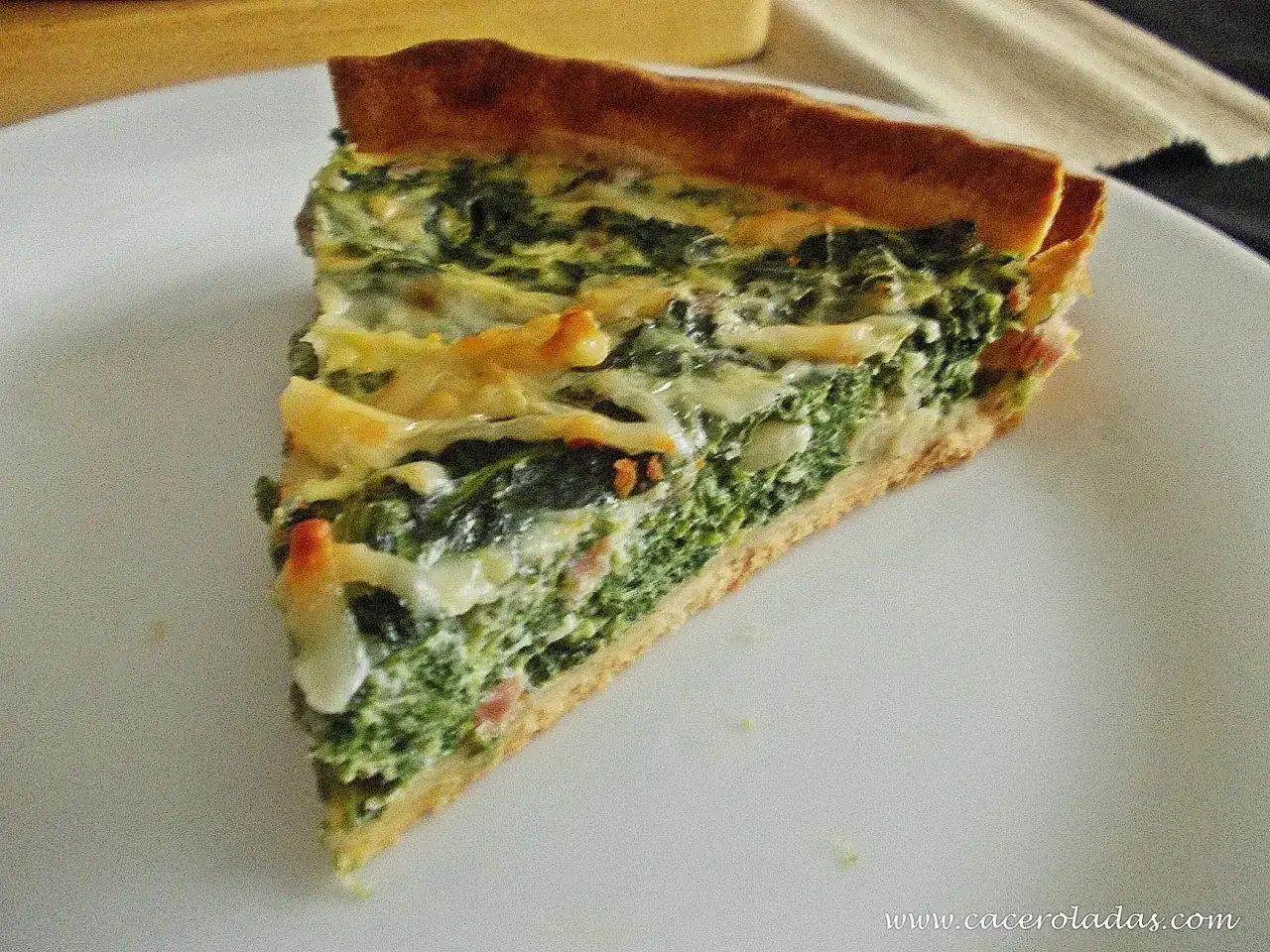 Rebanada de quiche de espinacas con queso derretido y trocitos de tocino, servida en un plato blanco.