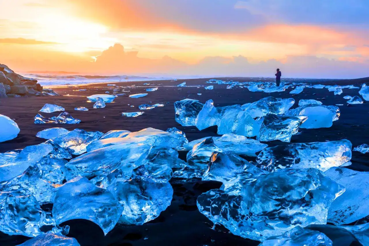 Fragmentos de hielo brillan como diamantes en la playa negra de Islandia, bajo un cielo anaranjado y azul.