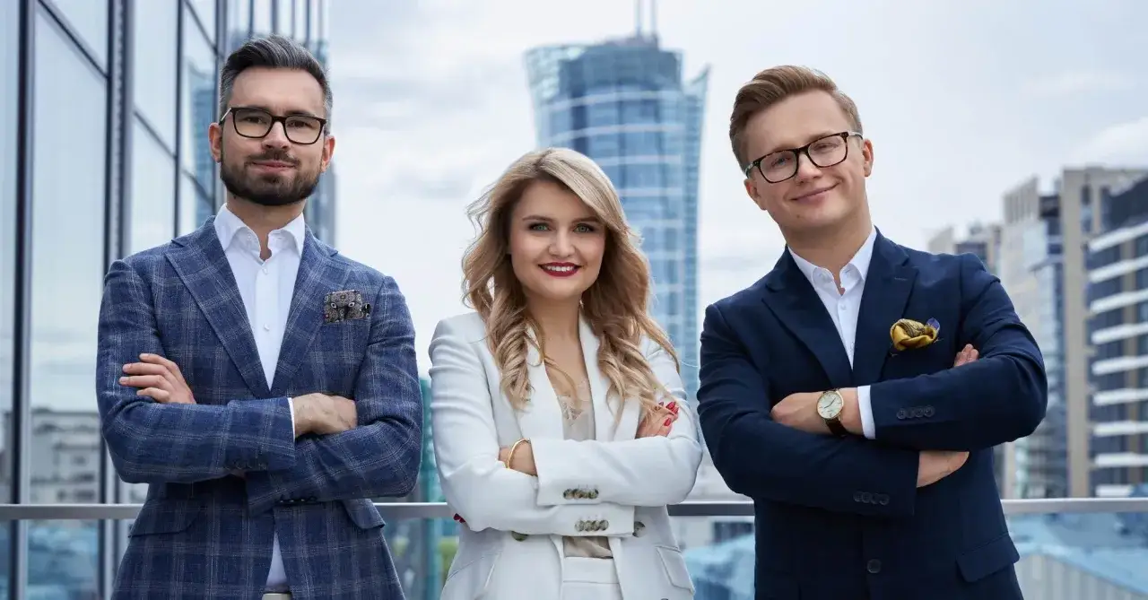 Trzy osoby w eleganckich garniturach stoją na tle nowoczesnych budynków.