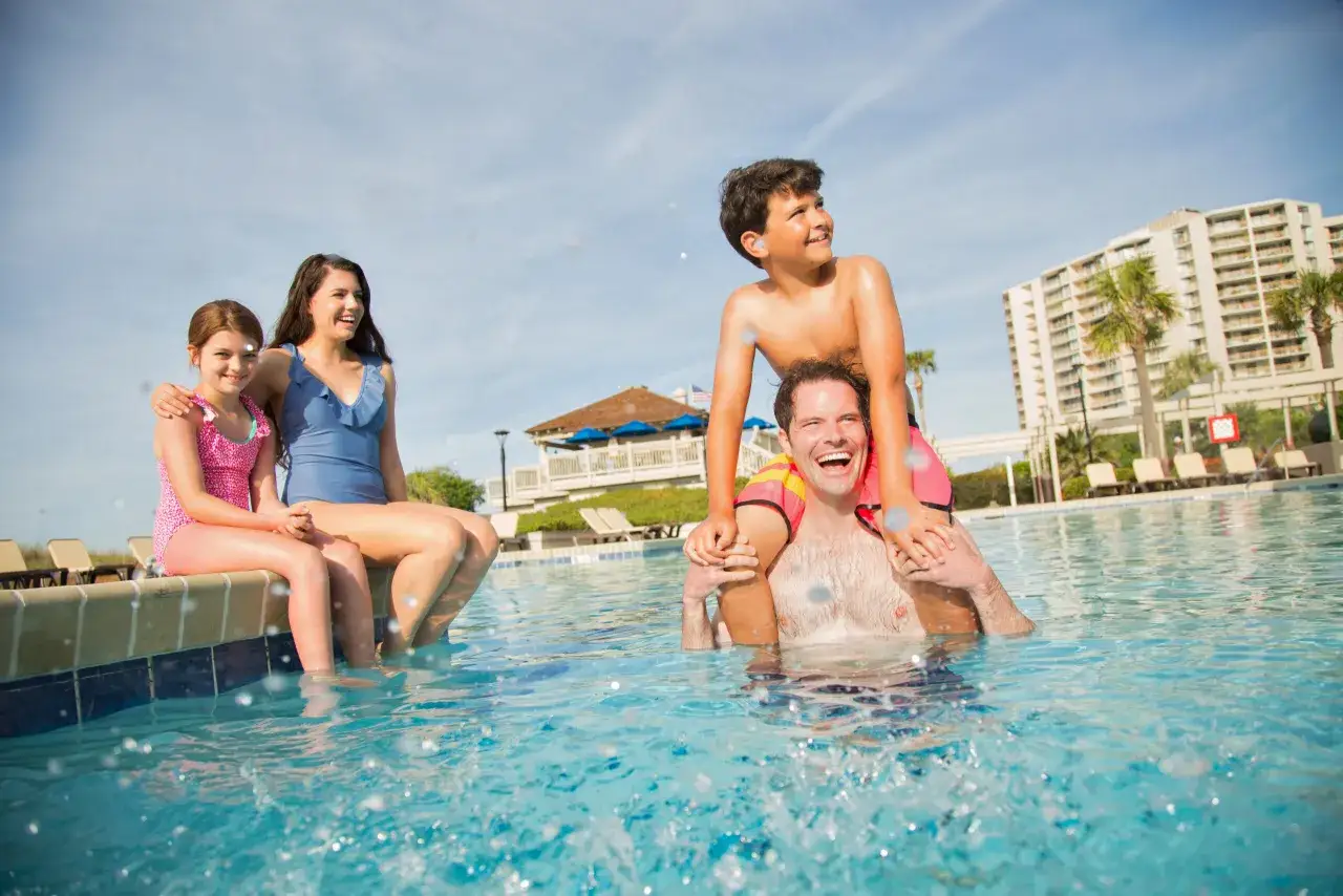 Rodzina cieszy się basenem przy Selectum Family Resort Side. Ojciec niesie syna na barana, a c&oacute;rki siedzą na brzegu.