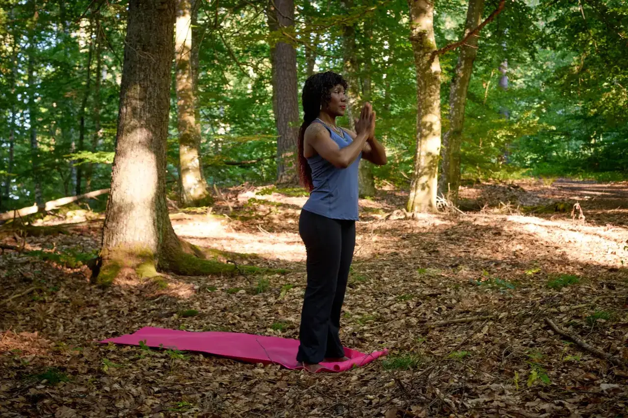 Kvinna med händerna i böneställning på en yogamatta i skogen, söker lugn från kronisk stress.