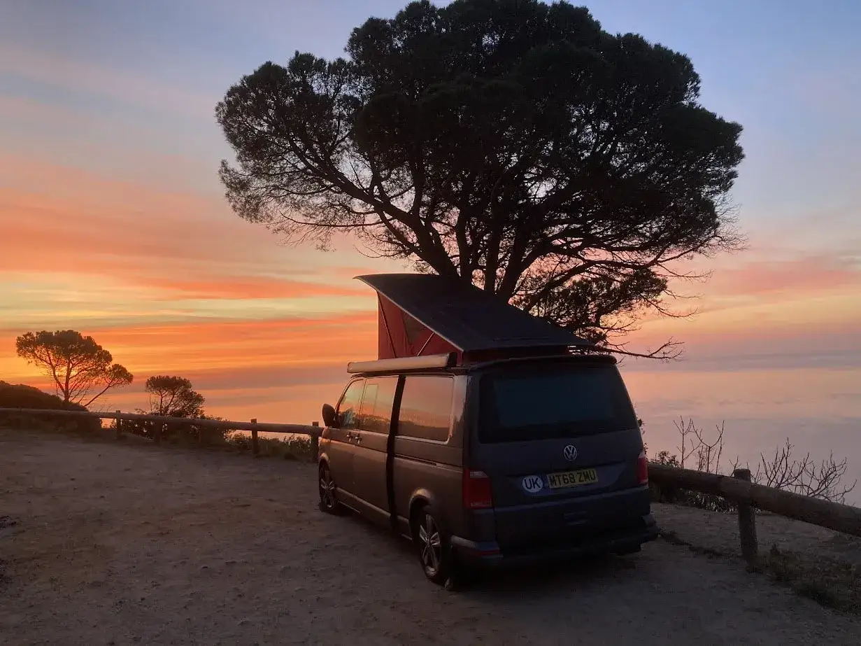 Un camping-car Volkswagen, prêt pour l'aventure en Angleterre, est garé sous un grand arbre au coucher du soleil. Le ciel est peint de couleurs vives.