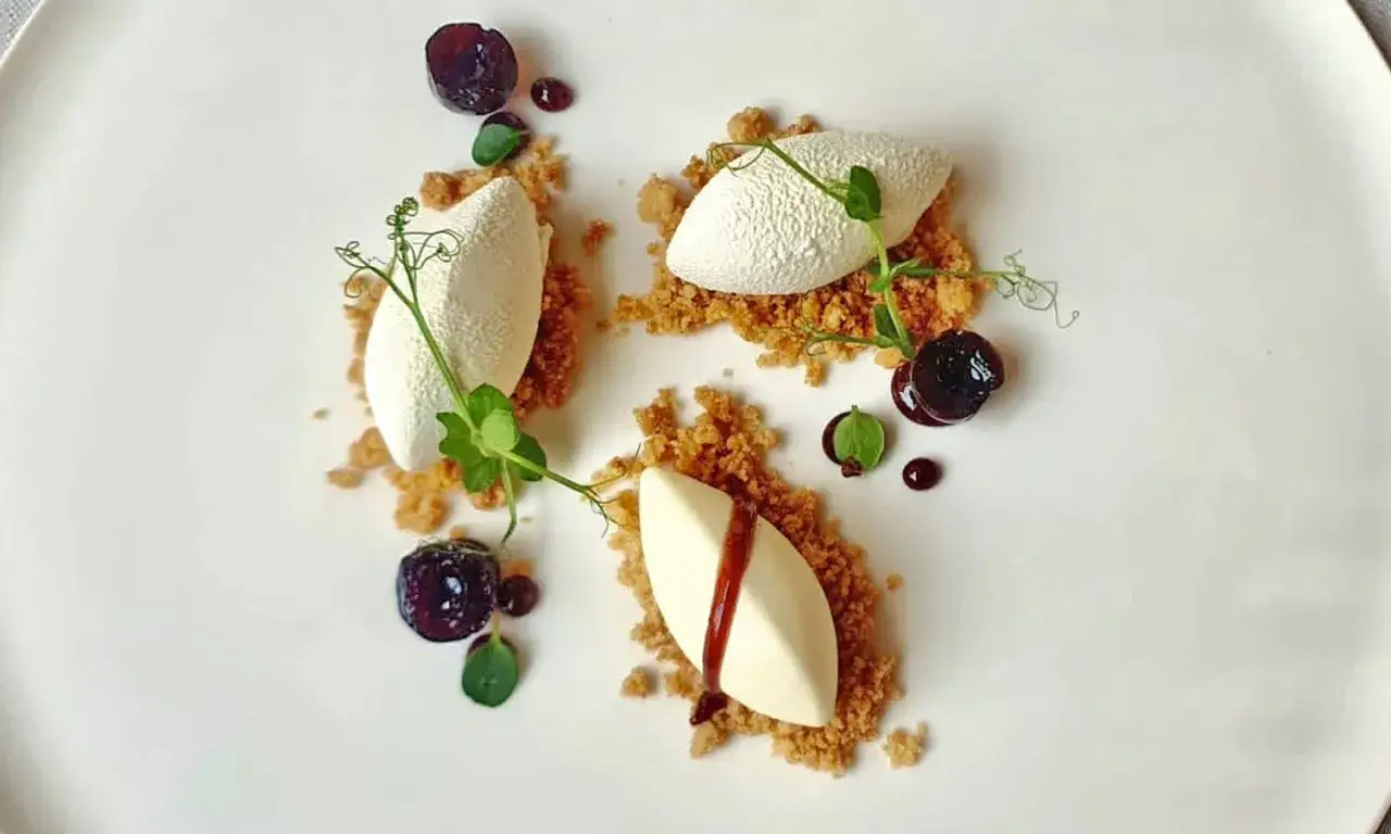 Delicata mousse di ricotta, guarnita con ciliegie sciroppate, briciole croccanti e germogli verdi su un piatto bianco.