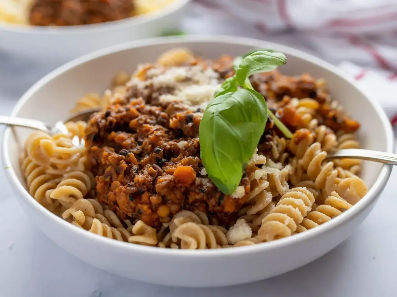 Makaron fusilli z sosem bolognese z soczewicy, posypany parmezanem i udekorowany listkiem bazylii. Idealny sposób, jak gotować soczewicę na obiad.