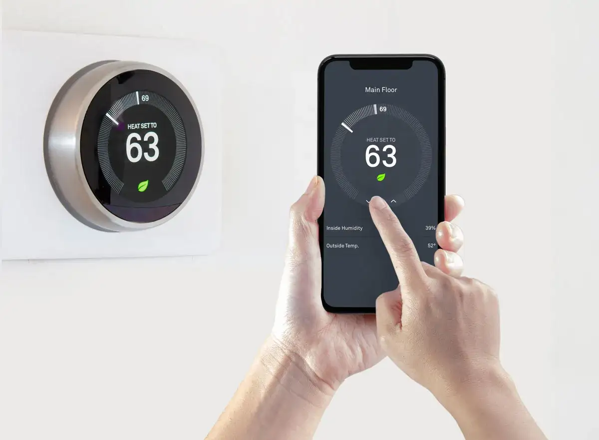 Une main utilise un smartphone pour piloter sa climatisation à distance. L'écran affiche la température intérieure et extérieure.
