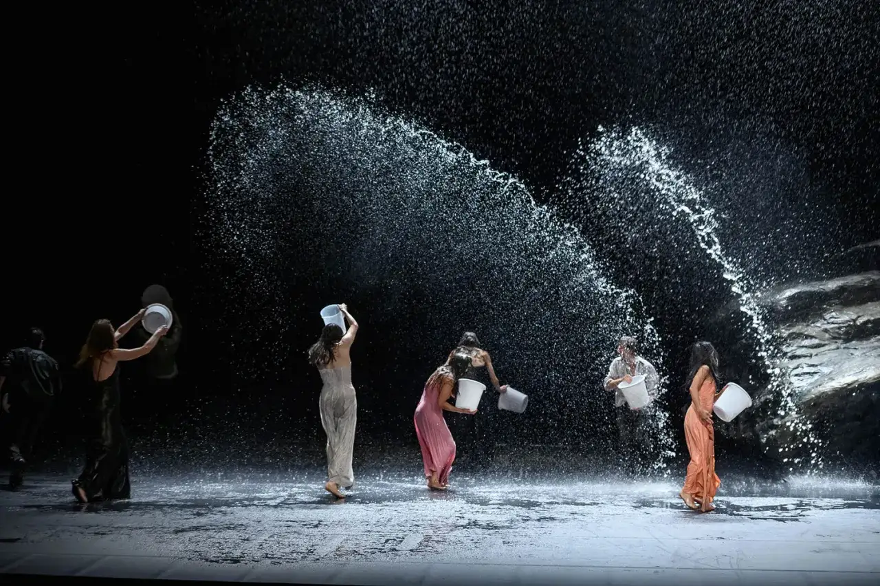 Tanztheater Wuppertal: T&auml;nzerinnen und T&auml;nzer sch&uuml;tten Eimer mit Wasser aus, w&auml;hrend ein Wasservorhang herabf&auml;llt.
