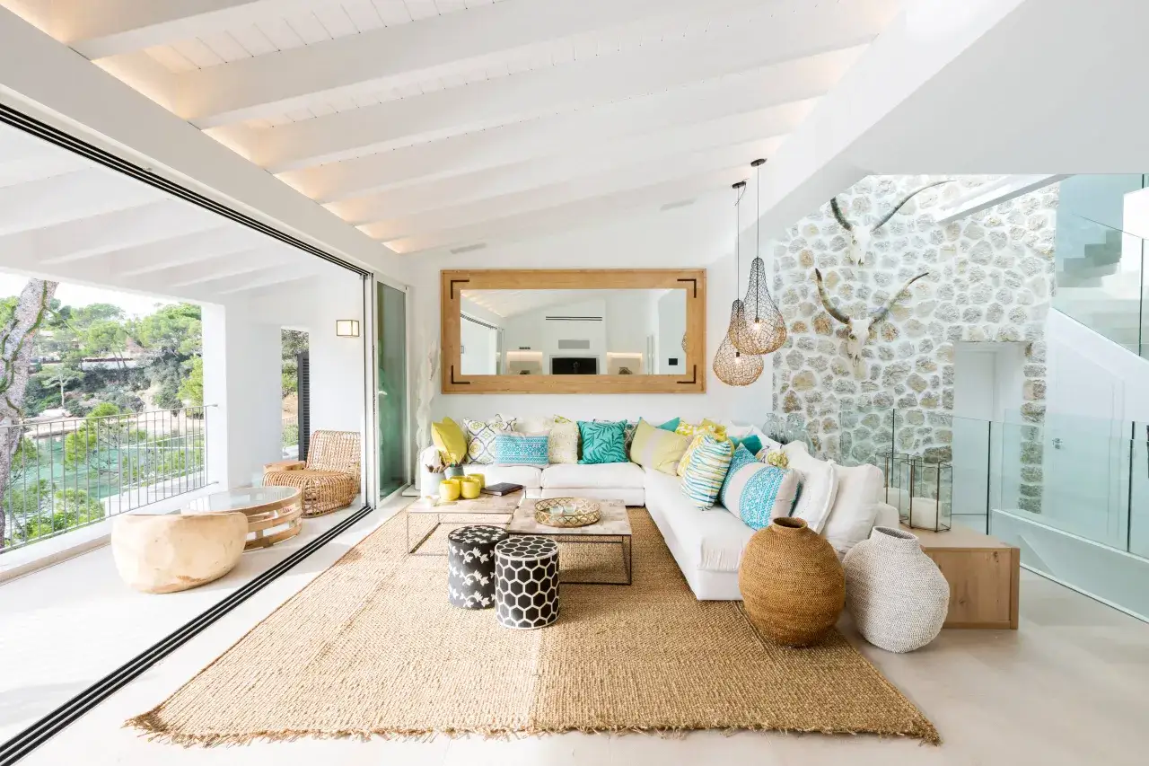 Helles Wohnzimmer mit Finca Style Einrichtung, wei&szlig;em Sofa, bunten Kissen, Jute-Teppich und Blick auf das Meer.