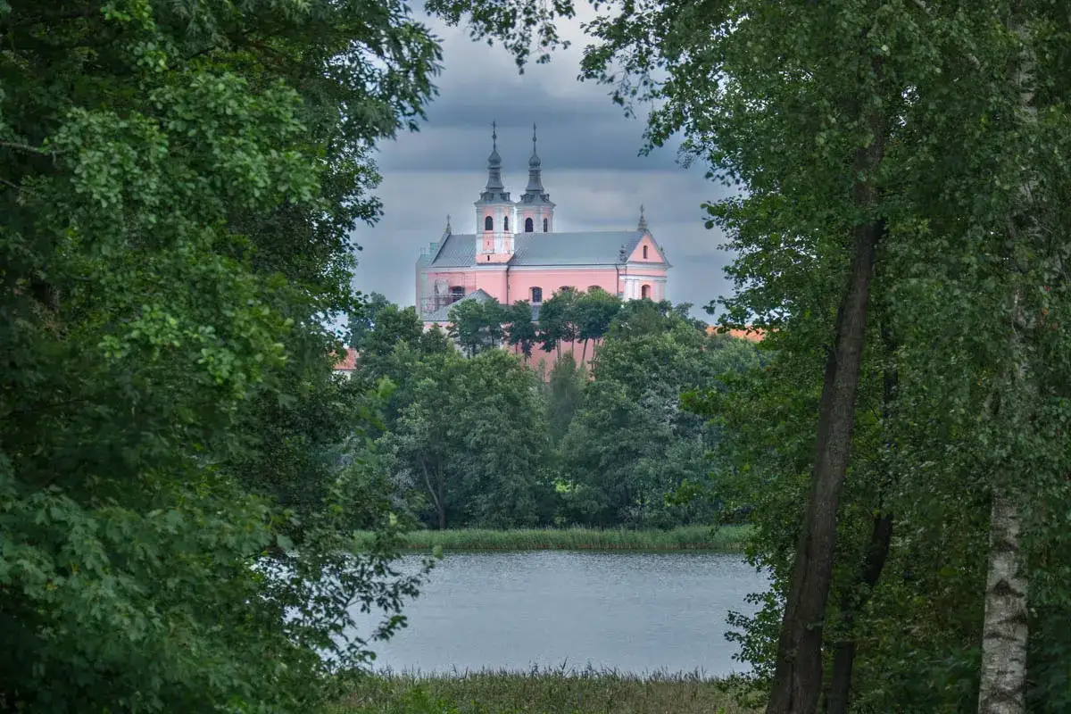 Różowy kościół z wieżami, jedna z atrakcji Wigierskiego Parku Narodowego, wyłania się zza zieleni drzew nad jeziorem.