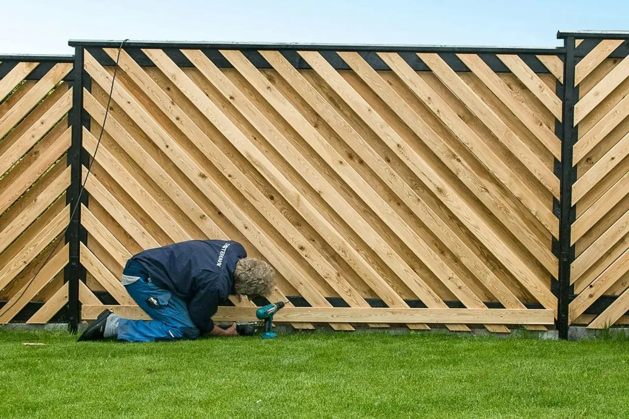 Man bygger plank med snedst&auml;llda tr&auml;ribbor och svarta stolpar. Gr&auml;smatta och bl&aring; himmel syns.