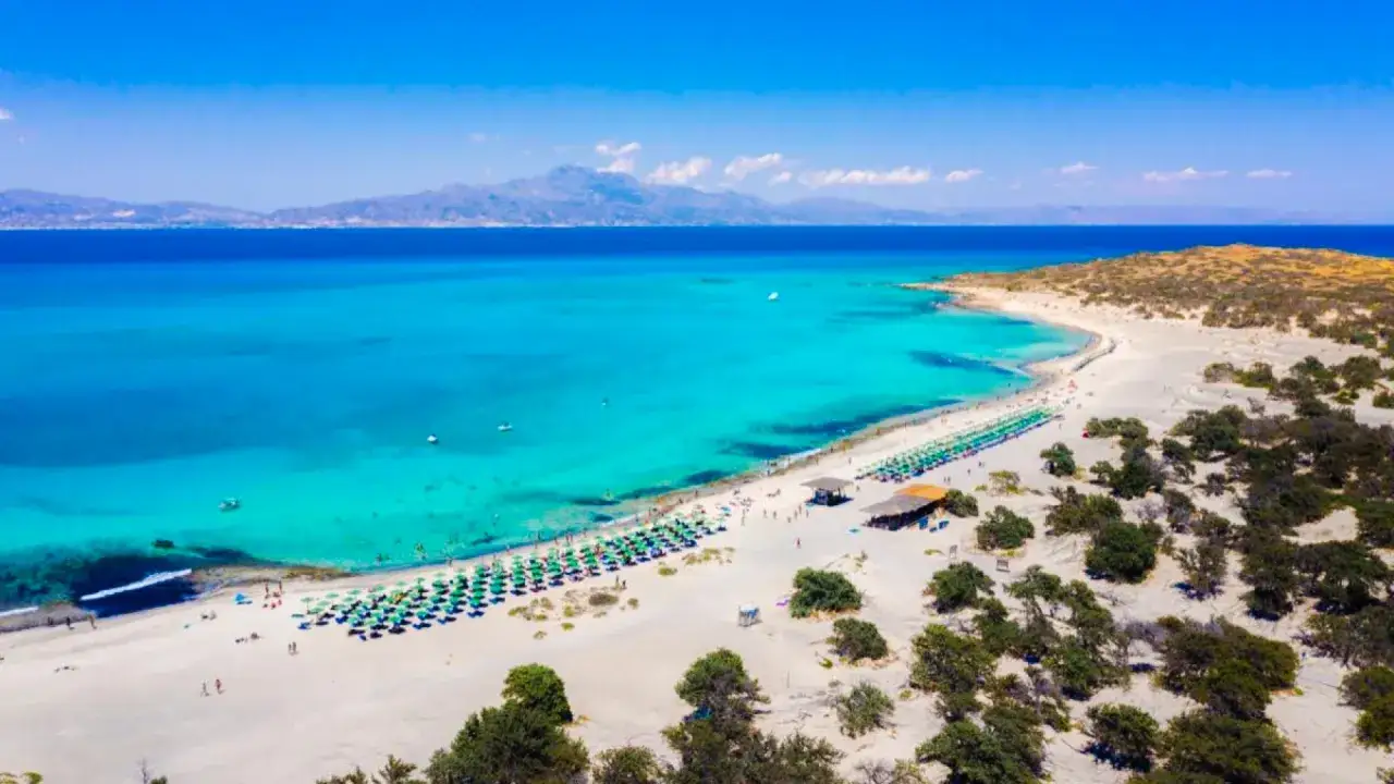 Rajska plaża Voulisma z turkusową wodą, białym piaskiem i rzędami zielonych parasoli.