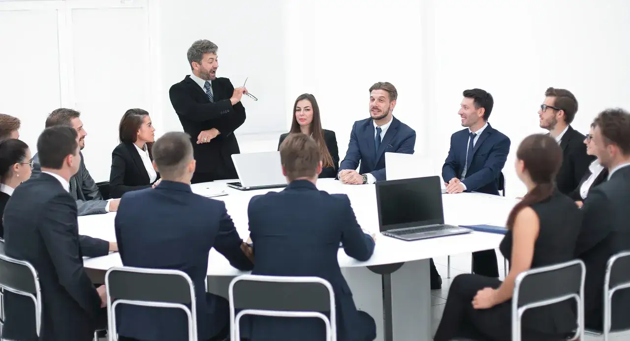 Mężczyzna przemawia na walnym zgromadzeniu, wskazując na coś. Wszyscy siedzą przy okrągłym stole, skupieni.