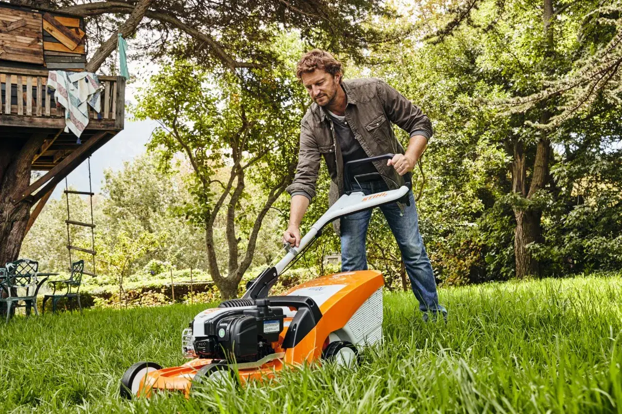 Mężczyzna z kosiarką STIHL, która jest symbolem ssania i mocy, pielęgnuje trawnik. W tle dom na drzewie.