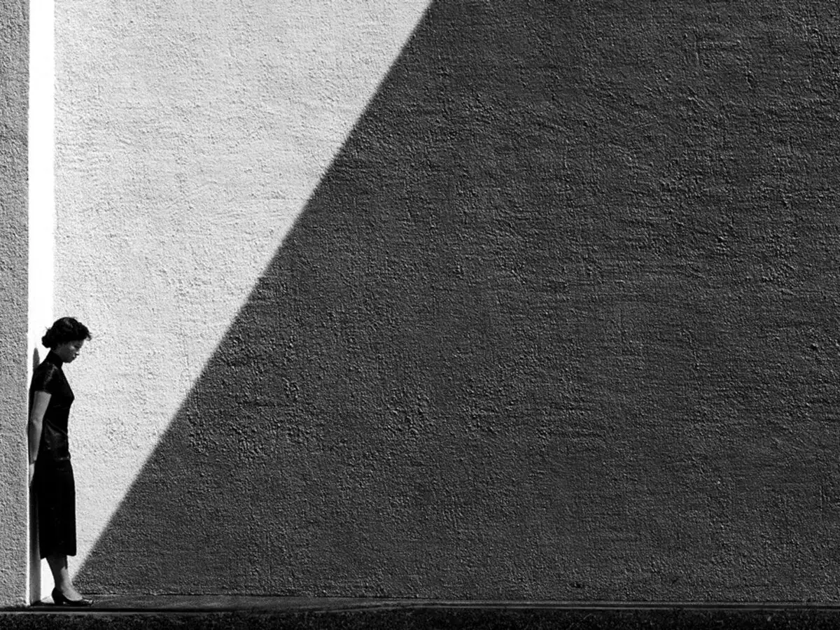 Donna in abito scuro, china, si appoggia a un muro diviso da un'ombra netta. Un fan ho.