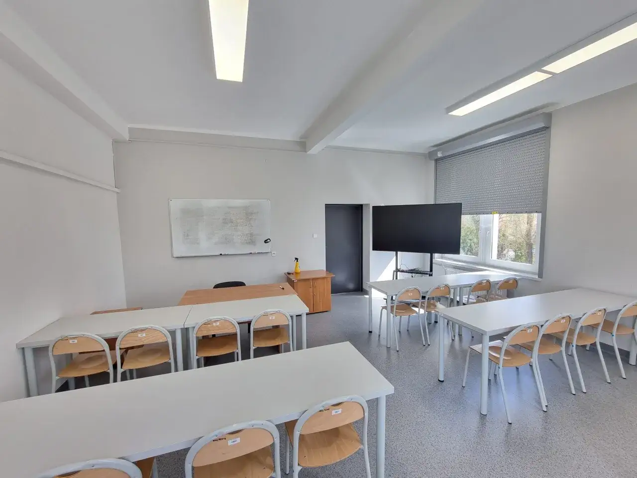 Nowoczesna sala wykładowa, idealna na studia w Raciborzu. Białe biurka, krzesła i tablica interaktywna tworzą przestrzeń do nauki.