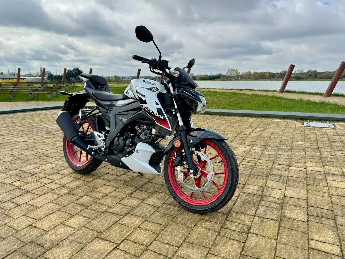 Biało-czarny Suzuki 125 szosowo-turystyczny z czerwonymi felgami stoi na wybrukowanym placu nad jeziorem.
