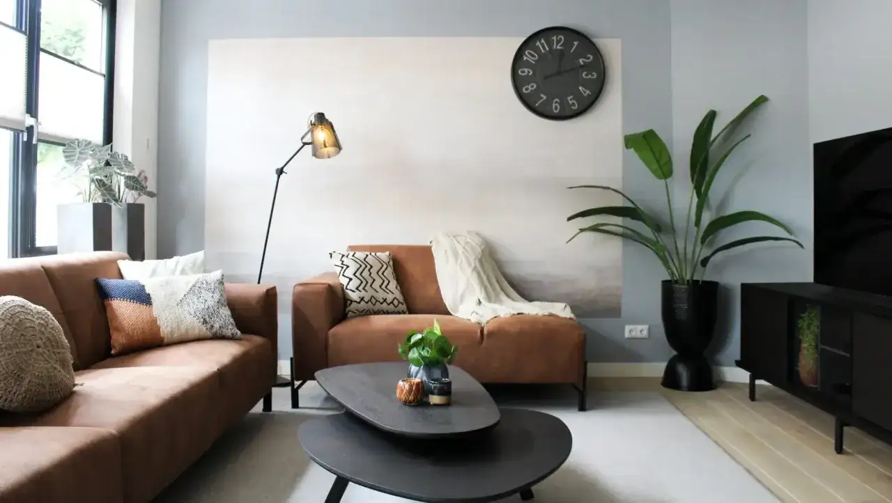 Quel budget pour meubler une maison neuve ? Salon moderne avec canapé marron, coussins décoratifs, table basse, plante verte et horloge murale.