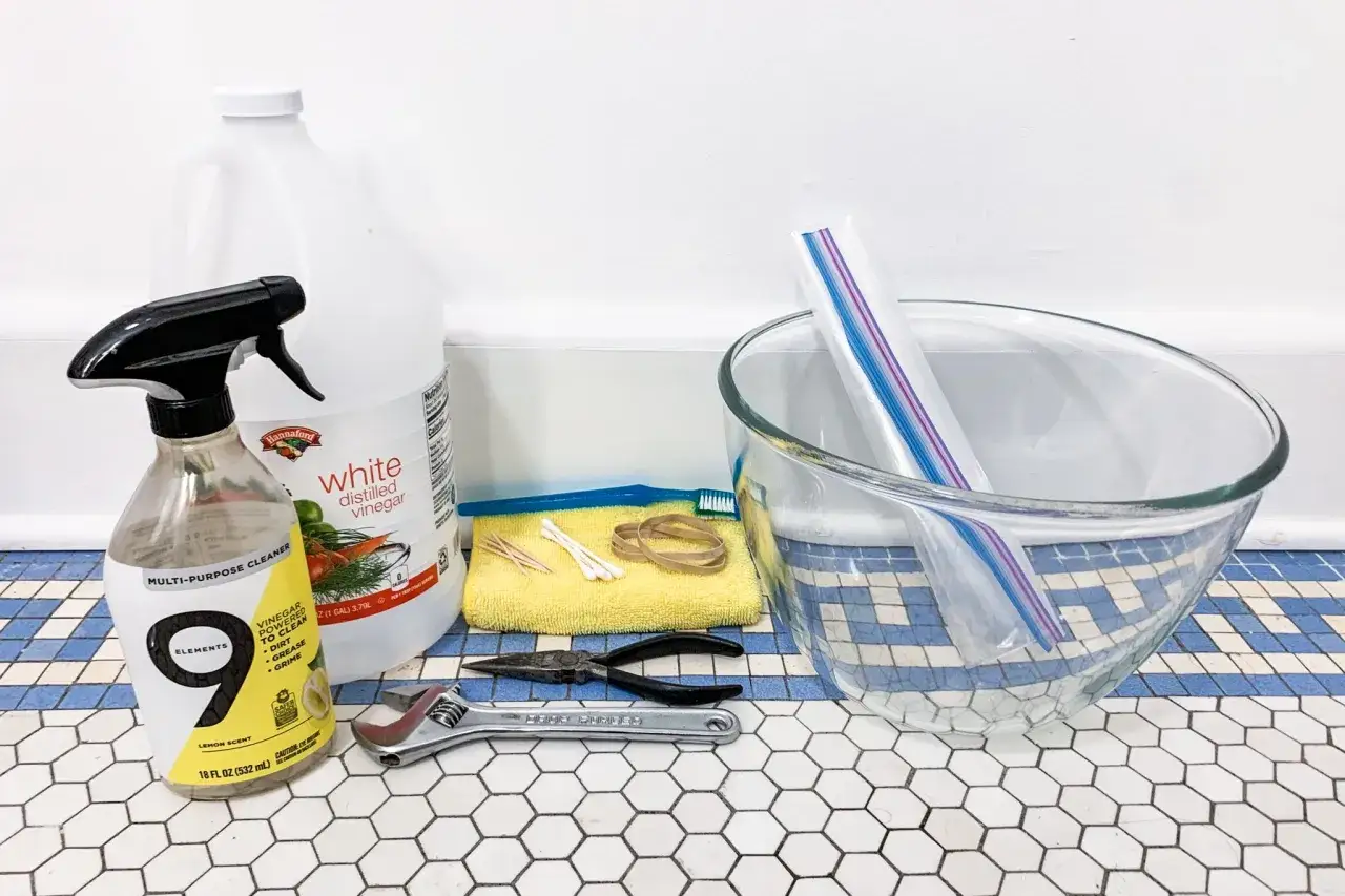 Gather supplies to clean a shower head with vinegar: distilled white vinegar, a spray bottle, a bowl, a plastic bag, a toothbrush, and tools.