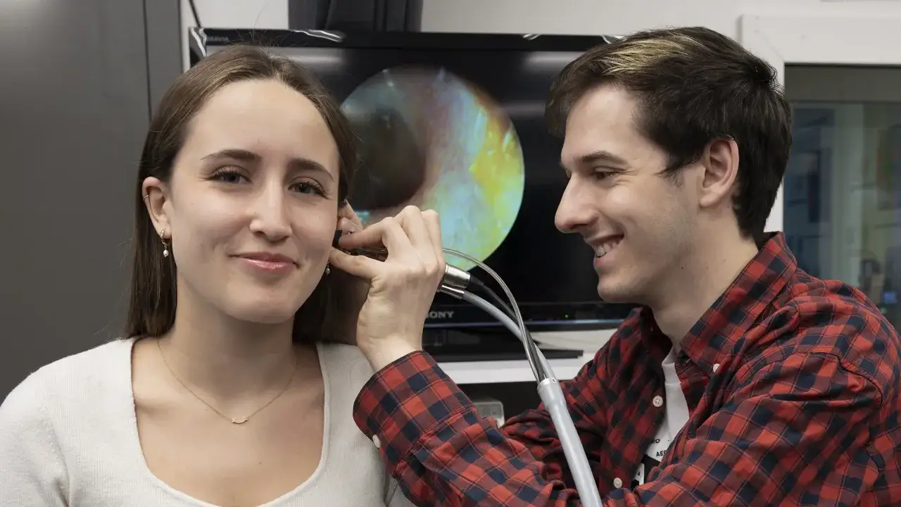 Un étudiant en formation audioprothésiste en Espagne à distance examine l'oreille d'une patiente, avec une image de l'intérieur de l'oreille affichée sur un écran.