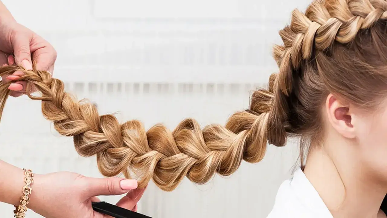 Eine Hand flechtet kunstvoll **geflochtene Haare** zu einem dicken Zopf. Die blonde Mähne glänzt im Licht.
