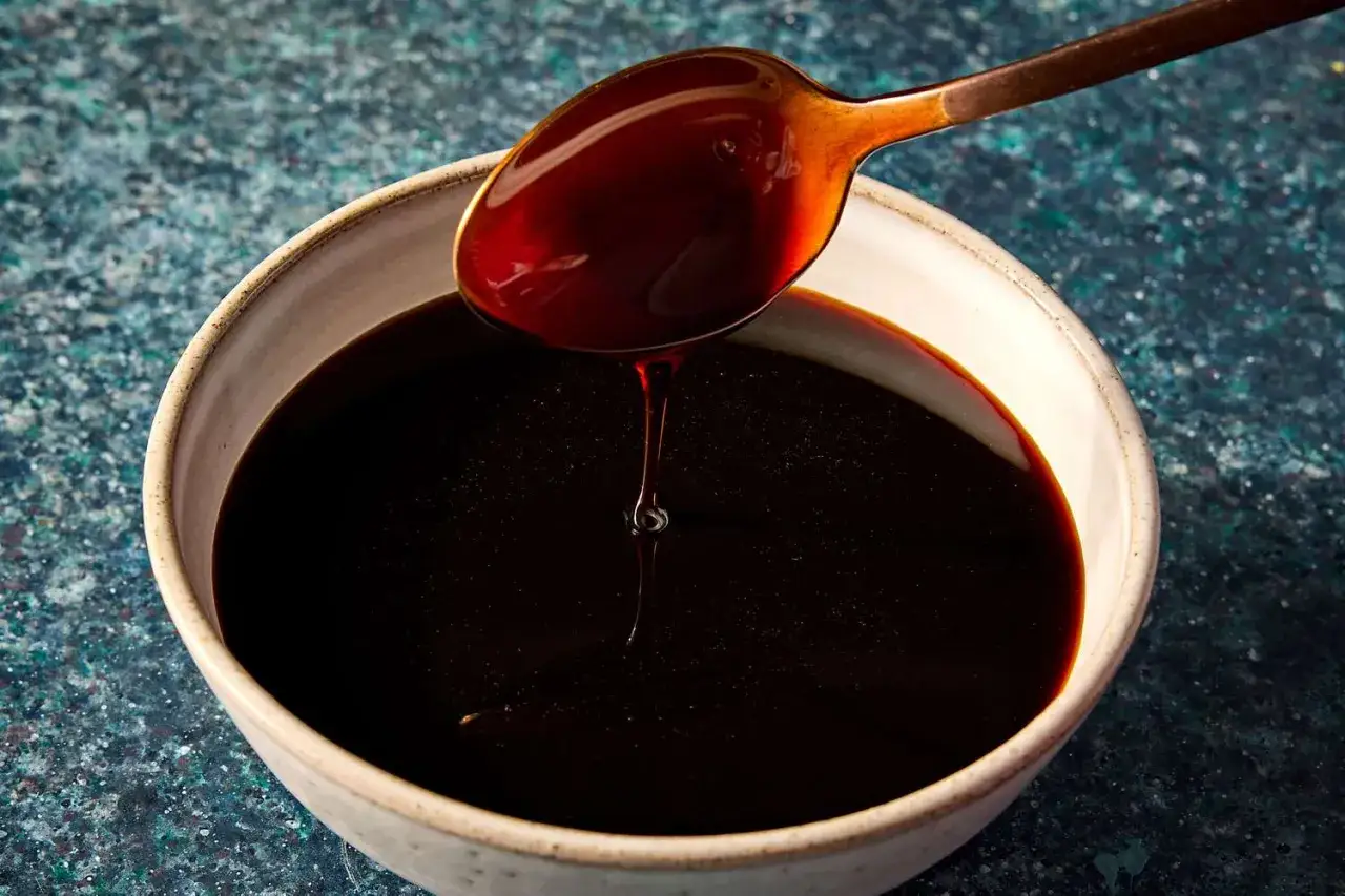 A spoon drips thick, dark liquid into a bowl, showcasing how to make sweet soy sauce.