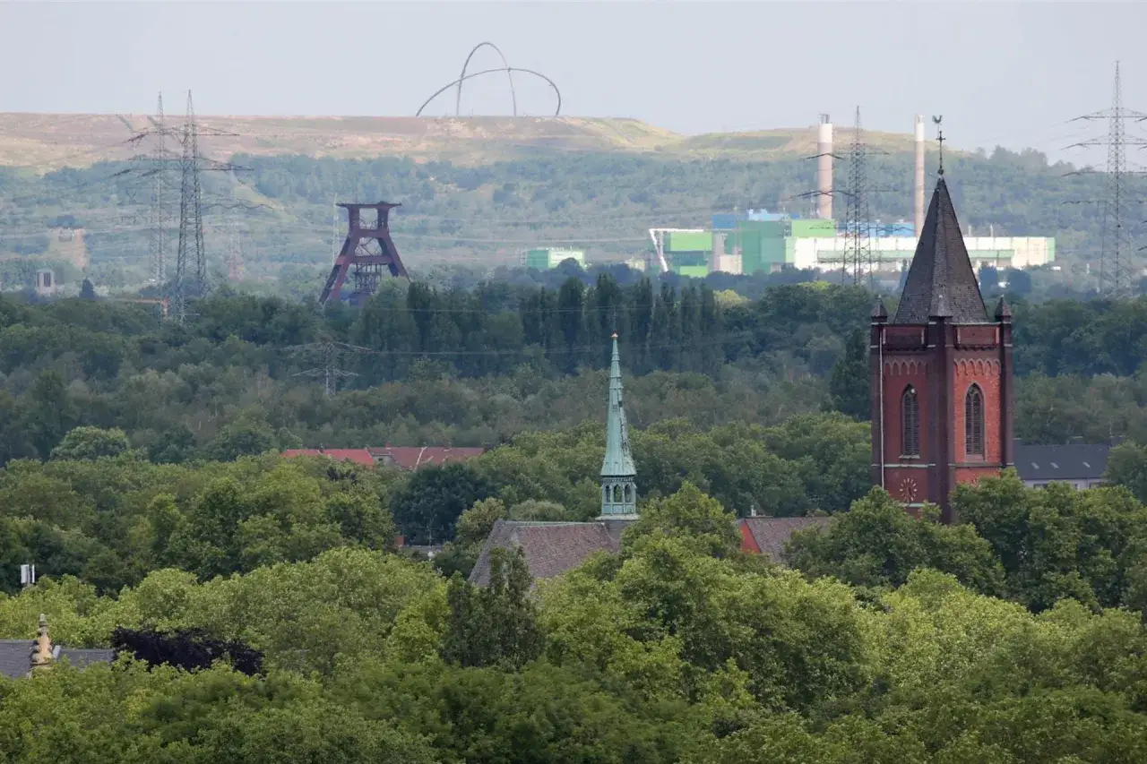 Kirchturm ragt aus Bäumen, im Hintergrund die Zeche Ewald Fortsetzung mit Förderturm und Industriebauten.