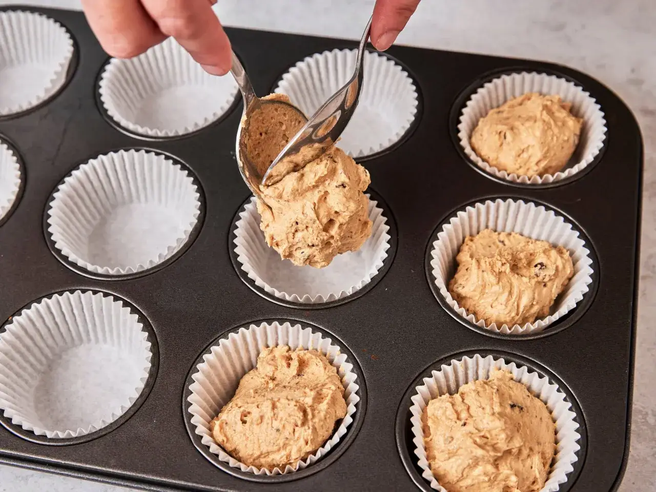 H&auml;nde verteilen Teig mit L&ouml;ffeln in Muffinformen. Das ist der Beginn vom Muffins backen.