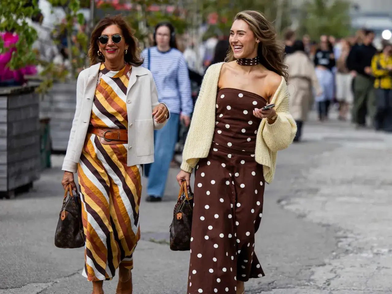 Zwei Frauen spazieren. Eine trägt ein gestreiftes Kleid, die andere ein braunes Kleid mit Tupfen. Beide sind stilvoll gekleidet und zeigen, wie man ein braunes Kleid kombinieren kann.