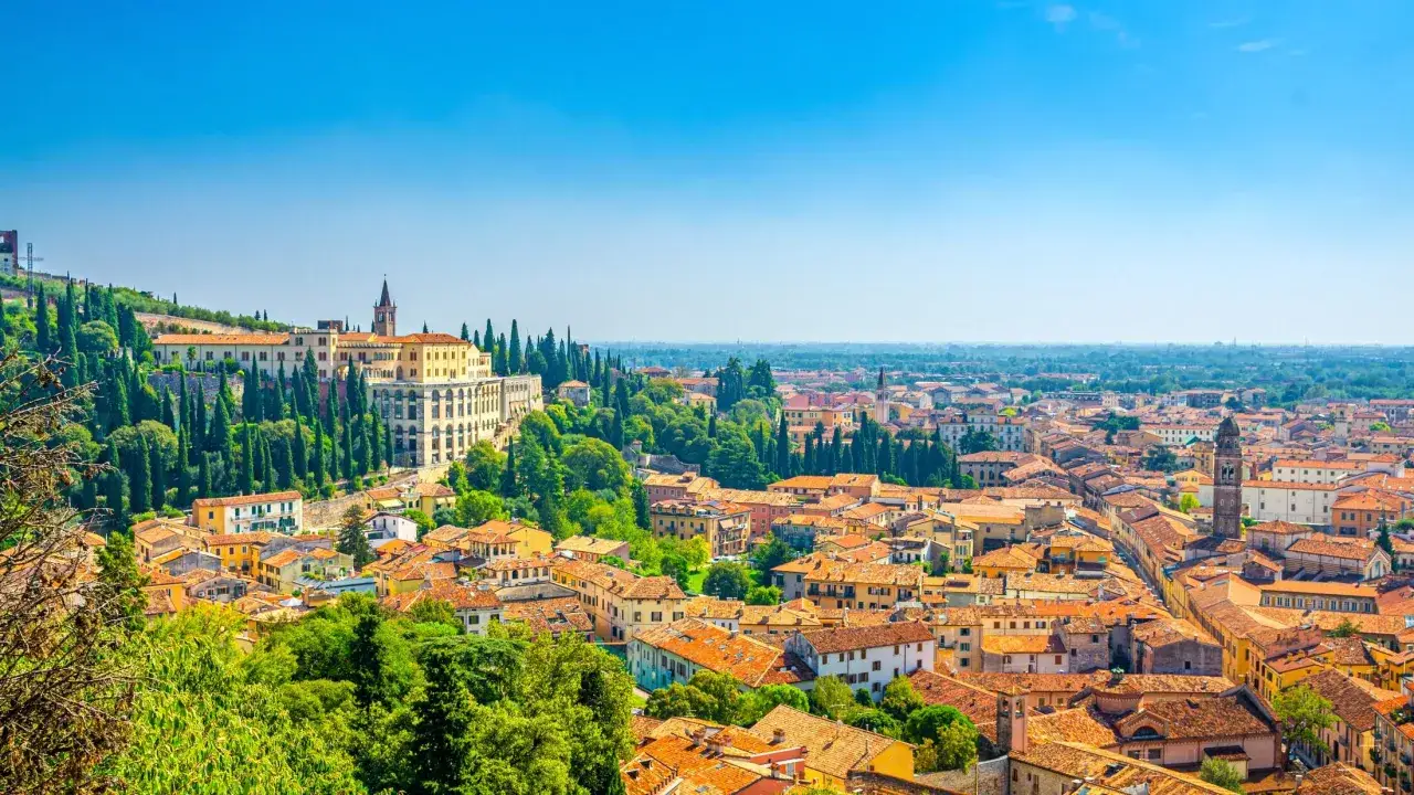 Vy över takåsar och grönska i Verona, Italien. En stor byggnad reser sig på en kulle, omgiven av cypresser.