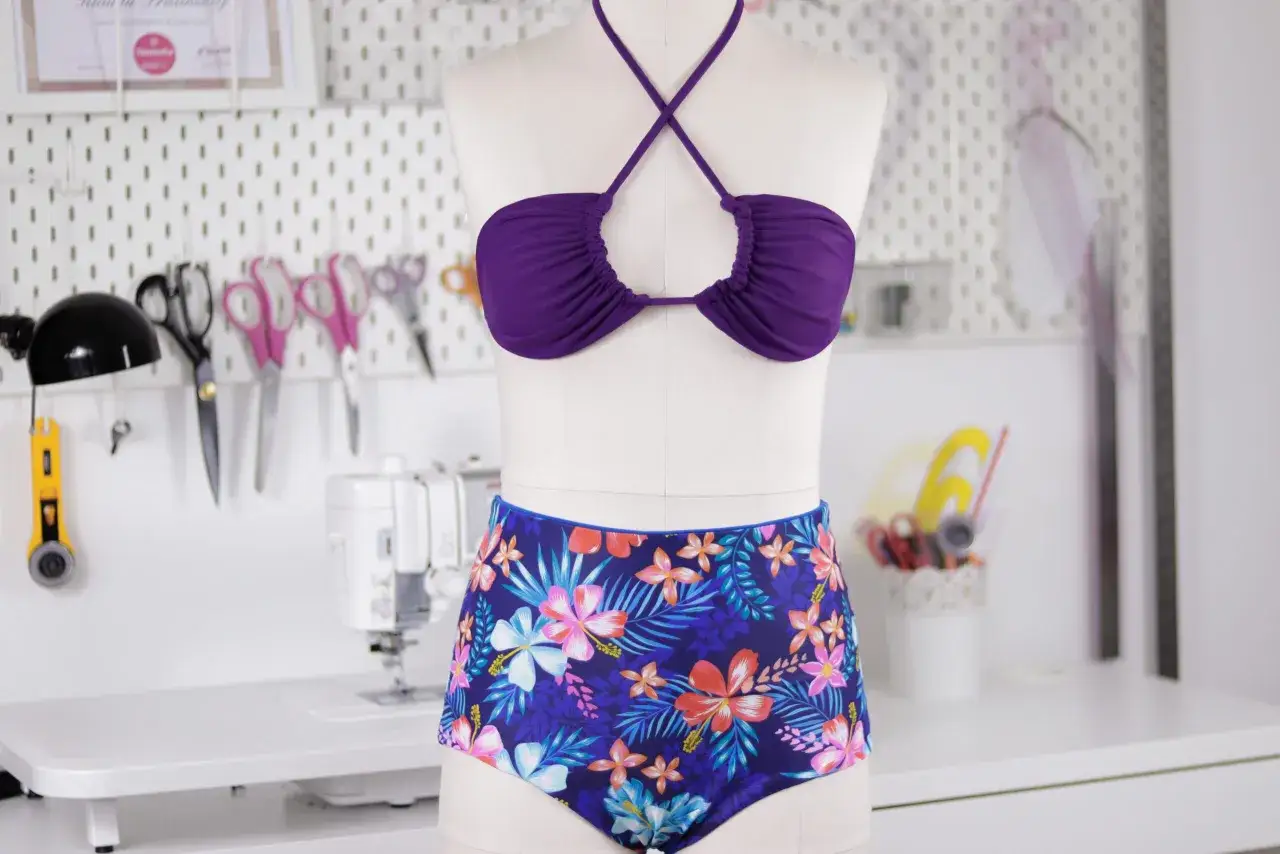 Jak uszyć strój kąpielowy? Fioletowy top bikini i figi w tropikalne wzory na manekinie, obok maszyna do szycia i akcesoria krawieckie.