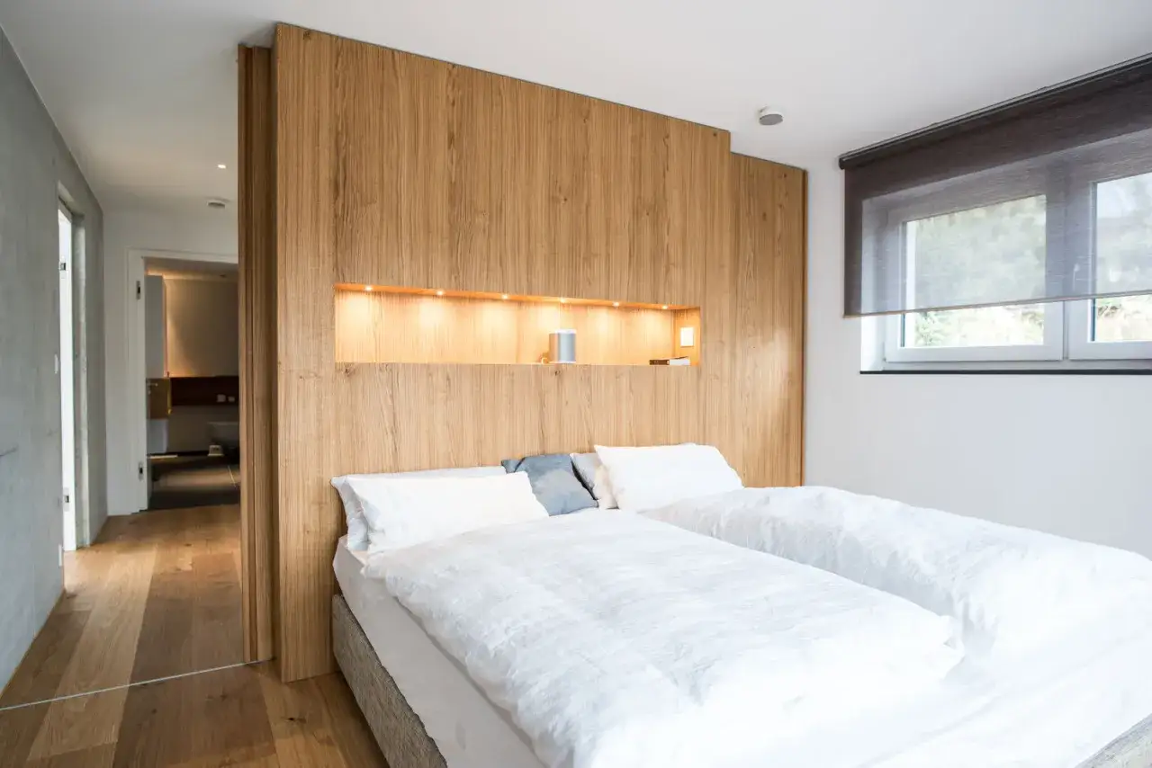 Ein moderner **Raumteiler vorm Bett** aus Holz trennt den Schlafbereich vom angrenzenden Bad. Zwei weiße Betten sind mit Bettwäsche bezogen.